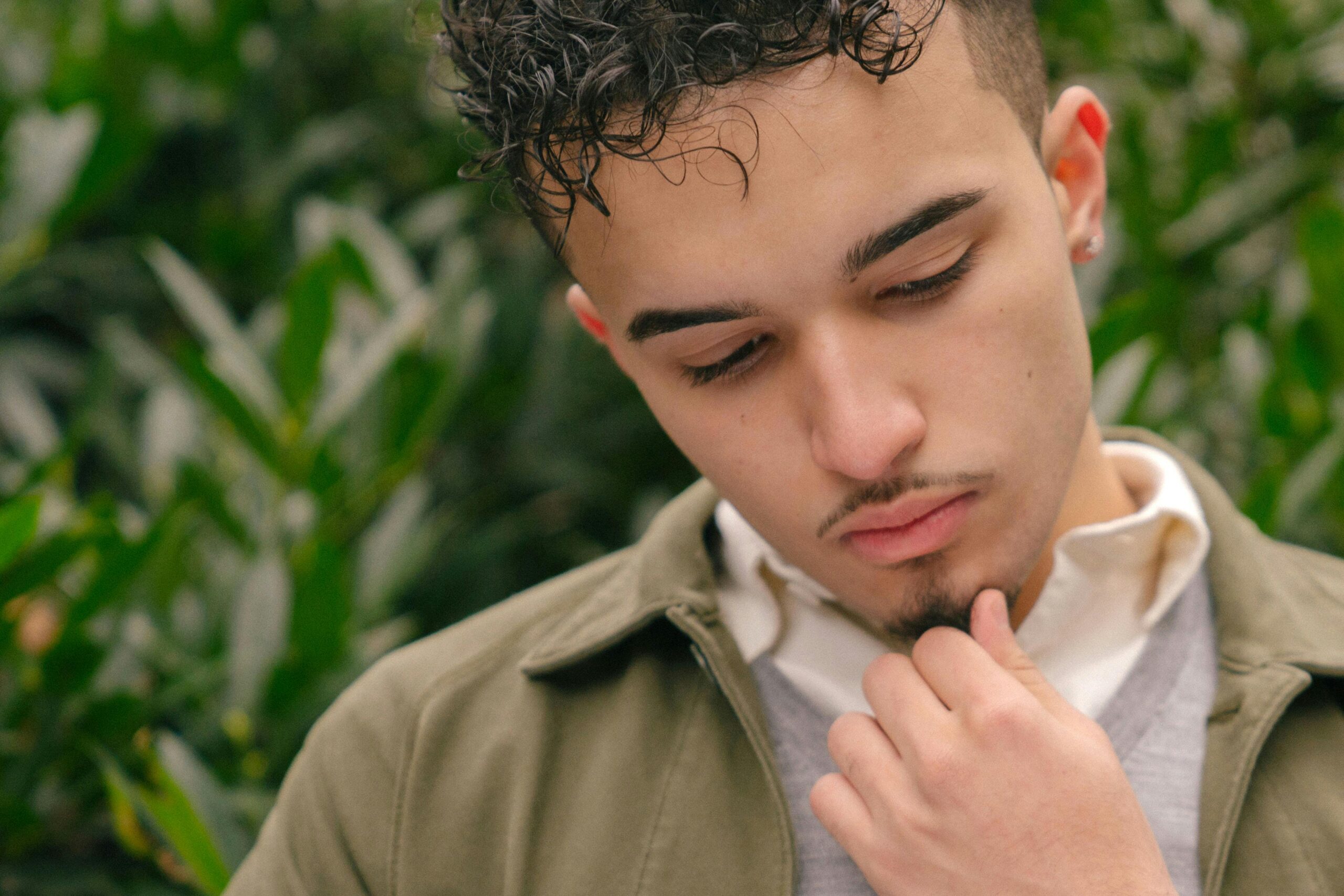 A young man with curly hair is deep in thought, standing outdoors with greenery in the background.