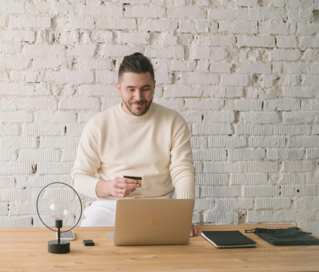 Adult man using credit card and laptop for online shopping in bright, minimalist office setting.