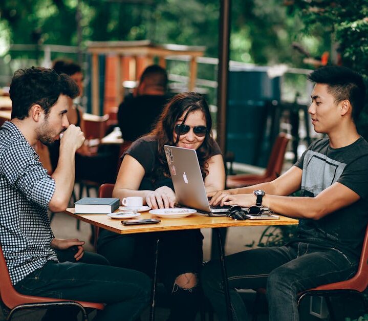 A group of young adults working on a laptop at an outdoor coffee shop, enjoying teamwork and collaboration.