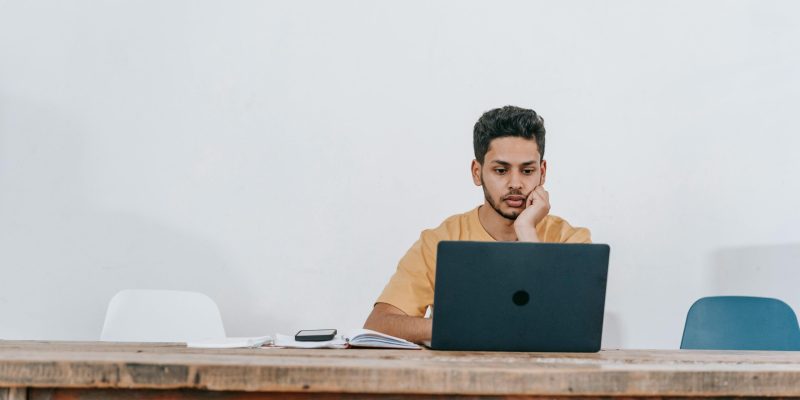 Focused young man studying on a laptop in a minimalist indoor workspace.